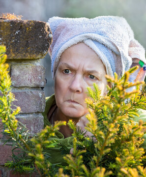 Eine Frau mit Kopftuch beobachtet und blickt neugierig. Sie steht versteckt zwischen einer Mauer und einem Gebüsch.
