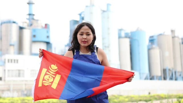 Sad asian female worker with mongolian flag standing in front of factory Worried asian girl in work clothes with flag of mongolia standing in front of industrial. High quality 4k footage
