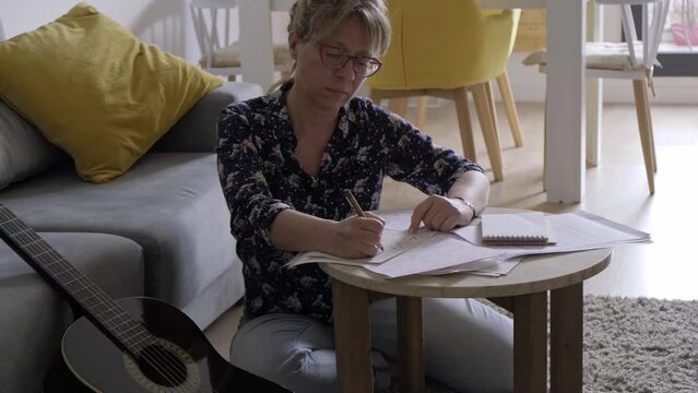 40-50 years old caucasian composer woman with guitar composes music and writes chords on paper , sitting on the floor of her house.
