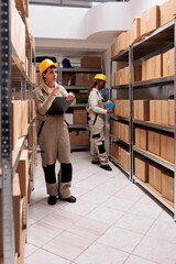 Warehouse manager in protective helmet counting boxes and writing on clipboard. Diverse stockroom employees working in storehouse, monitoring shipping process and packing parcels with adhesive tape