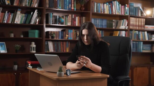 The girl works on her laptop and types on it. She gets a phone call and interrupts her work to answer it. After the call, she writes some information in her notebook.
