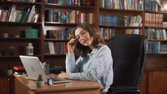 A dreamy young woman, surrounded by many books, is enjoying her time sitting at a desk with a laptop, with some restlessness in her soul and thoughts wandering in her head.