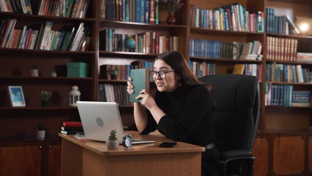 Sitting at the table, the girl is having an online conversation through a webcam, emotionally showcasing a book, and in the end, money falls from above.