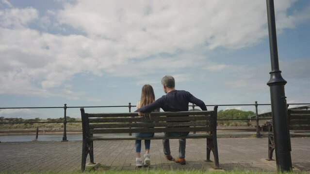 Dad and daughter sit together on bench chatting and cuddling, wide shot