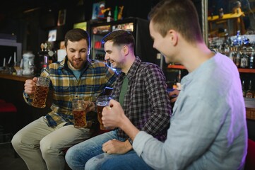 Three young men in casual clothes are smiling, holding bottles of beer while standing near bar counter in pub