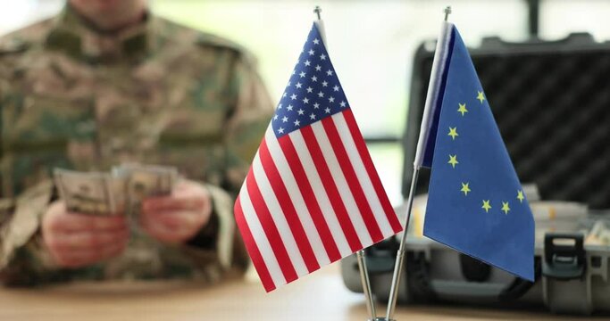 American and European Union flags against military officer counting money as international assistance. Man in military uniform sits at table in office slow motion