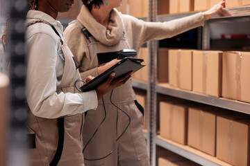 Women wearing overalls scanning parcels in ecommerce retail store warehouse. Post office storage employees searching customer order package, checking cardboard boxes with bar code scanner and tablet