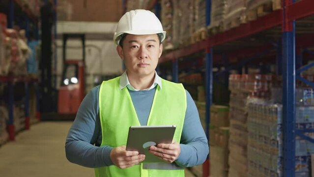 Competent male manager surfing internet on digital tablet while standing at storehouse with huge high shelves around. Asian man in reflective hardware checking supplies updating online info.
