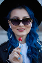 Portrait of a beautiful diverse woman with dyed blue hair licking a lollipop. Cheerful female person wearing a hipster hat, sunglasses and listening to music in wireless headphones