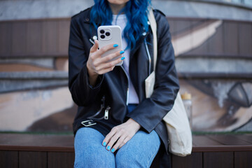 Fototapeta premium Young woman using a smartphone outdoor. Unrecognizable person sitting on a bench and browsing mobile app on a cellphone. Stylish female wearing black leather jacket, jeans and shopper bag