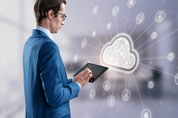 Attractive young european businessman using digital tablet with white cloud hologram and various icons on blurry office interior background. Cloud computing and storage server concept.