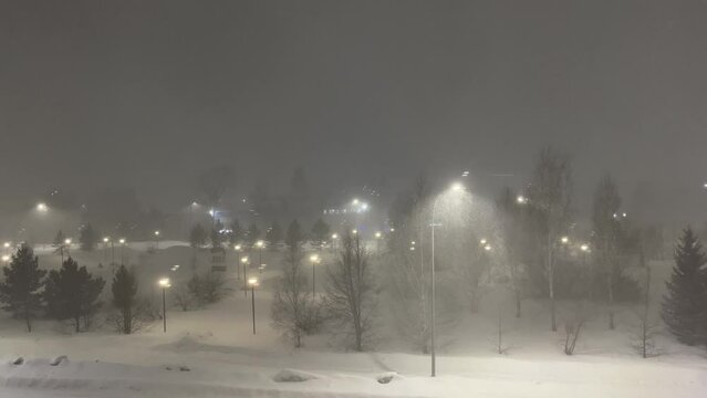 Snow storm in the night city. A view from above on the road along which the car is driving and a half-empty parking lot and a strong snow storm at night. High night lights light the way