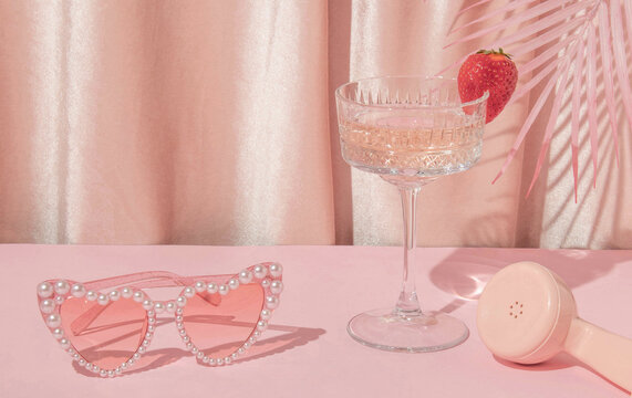 Summer creative layout with heart sunglasses, cocktail glass, phone handset and palm leaves with pastel pink curtain background. 80s or 90s retro aesthetic idea. Minimal summer fashion idea.