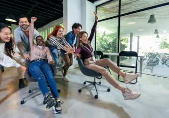 Young cheerful businesspeople in smart casual wear having fun while racing on office chairs and...