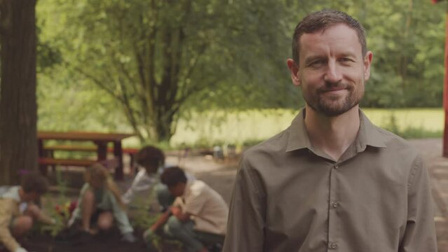 Waist up portrait of smiling Caucasian male ranger or eco activist posing for camera standing in summer public park while diverse school children planting together in blurred background