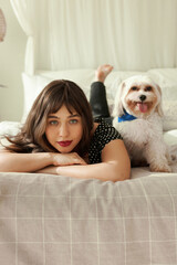Vertical portrait of pretty charming female with red lipstick on lips lying on bed with her white chinese dog pet next to her, looking at camera resting chin on hands, spending weekend at home