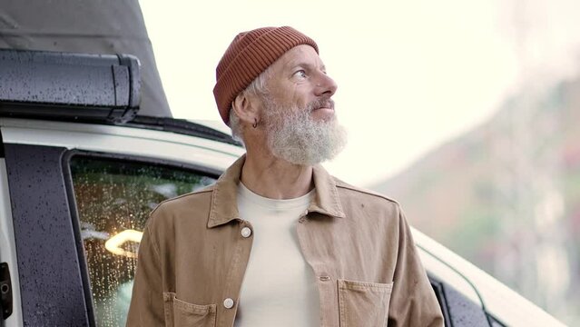 Active old happy hipster man standing near rv camper van on vacation. Mature traveler enjoying landscape view, looking away waking up in the morning feeling free in camping tourism nature park.