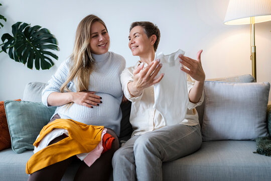 Smiling senior woman showing baby clothes to pregnant daughter sitting on sofa at home