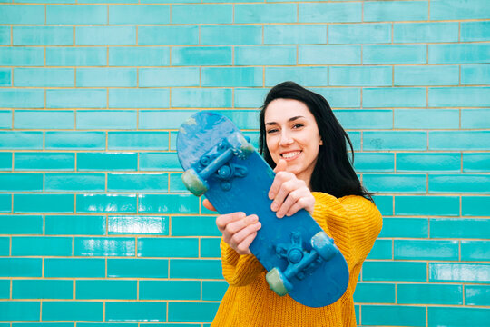 Smiling woman in sweater showing blue skateboard in front of brick wall