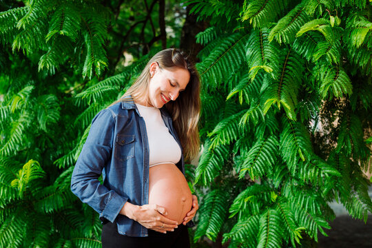 Smiling pregnant woman with hands on stomach standing in front of tree