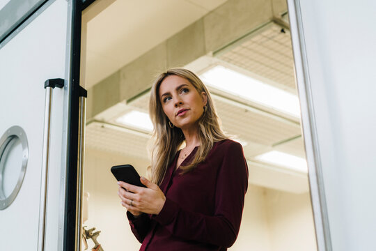 Thoughtful businesswoman holding mobile phone near doorway in factory
