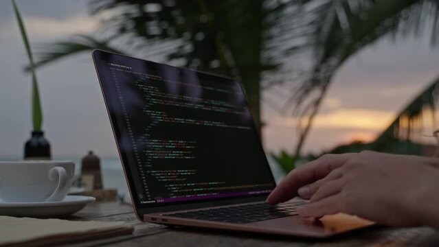 Young female developer working on laptop by the ocean at sunset. Woman freelancer coding at outdoor tropical cafe. Caucasian girl working remotely typing on computer at exotic location close-up