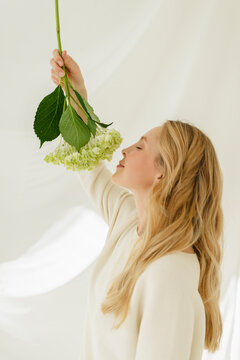 Blond woman smelling hydrangea by translucent curtain