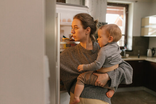 Mother carrying baby boy looking into refrigerator in kitchen at home