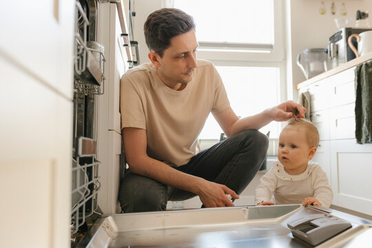 Cute baby girl and father loading utensils in dishwasher in kitchen at home
