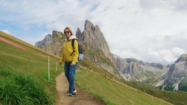 A blonde woman carrying a backpack is using a professional camera to capture photos of the breathtaking Seceda mountain.