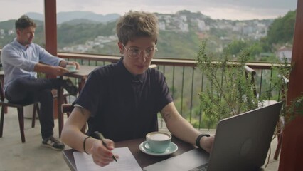 Medium shot of young businesswoman using laptop and taking notes while working remotely on terrace of cafe on mountain top, man drinking coffee in background