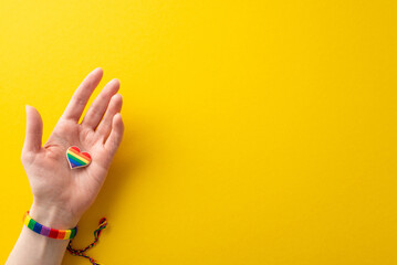 Observe LGBTQ History Month with this first person top view photo of female hand wearing a rainbow-colored bracelet and holding a heart-shaped pin badge on yellow background with space for text or ad