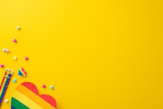 A top view flat lay of parade symbolic accessories, including wristlet, badge, hearts, rainbow card, on yellow backdrop with empty space for text or ad, celebrating the theme of Pride and inclusivity