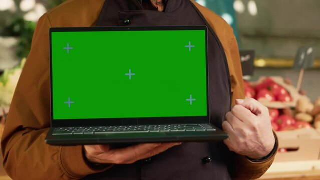 Senior seller showing greenscreen display on laptop, wearing apron at farmers market shop. Male vendor hoding pc with isoated chroma key and blank copyspace mockup template. Close up.