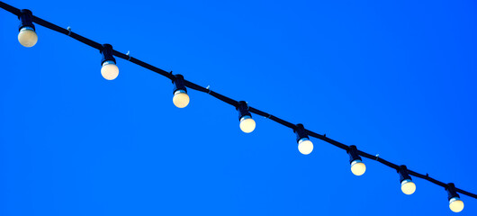 White electric light bulbs on a black power cord diagonally against the blue sky background