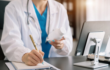 Pharmacist writing making notes in drug information using laptop computer sitting at desk. Woman physician, nurse or pharmacist wearing white coat writing in paper notebook.