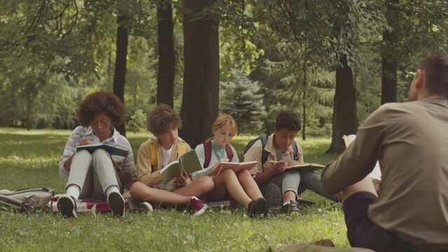 PAN shot of group of multiethnic school children and their male teacher having outdoor lesson on grass in park