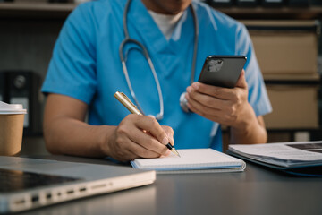 Physician or practitioner in room writing on blank notebook and work on laptop computer, smartphone on the desk,  Medic tech concept.