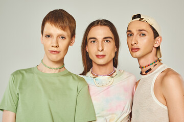 portrait of three lgbtq people with colorful beads looking at camera while standing together on grey background in studio, celebration of pride month concept