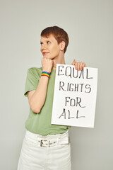 portrait of smiling queer activist wearing lgbt flag bracelet and holding placard with equal rights for all lettering looking away during pride month on grey background