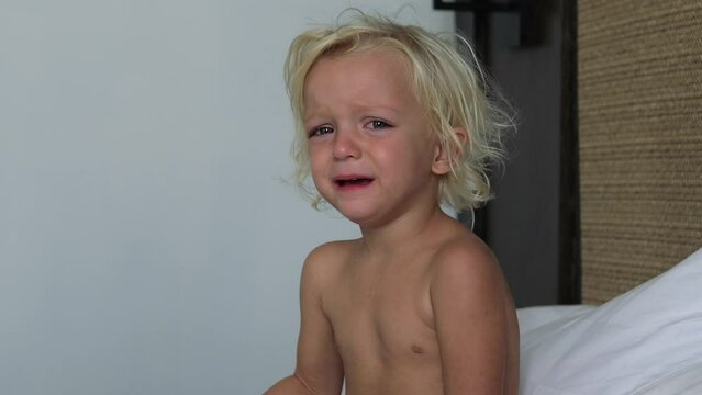 Naked little Caucasian blonde child is crying bitterly, sitting on the bed. Hysteria, grief in the life of a small child. The child is hysterical to get what he wants, to show his dissatisfaction.