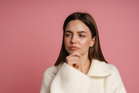Pensive woman touching her chin and looking away while standing isolated over pink wall