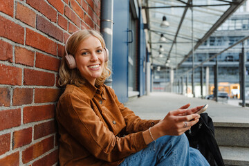Beautiful woman listening music with headphones and smartphone while sitting outdoors near brick wall