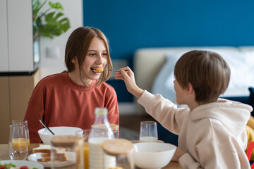 Happy kids siblings having fun together eating cereals in morning sitting at kitchen table, brother feeding sister corn flakes. European family starting day with healthy breakfast, enjoying meal
