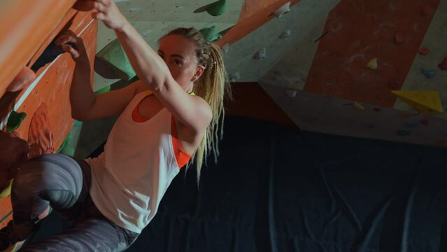 Hard training of a young pretty blonde girl or woman on an artificial climbing wall indoors. A female climber is thinking where to grab on to the wall of the climbing wall. High quality 4k footage