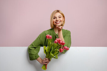 Happy young woman blowing a kiss while holding bunch of tulips against pink background