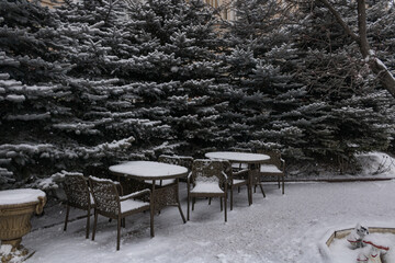 Plakat Snow-covered tables and chairs of the outdoor cafe next to the fir trees. An empty cafe. No people. Winter in the southern city of Russia.