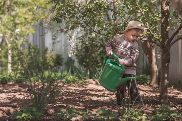 Naklejka premium A cute boy in a hat plants and waters seedlings from a watering can in a summer garden, outdoors. The concept of gardening and teaching a child to work.