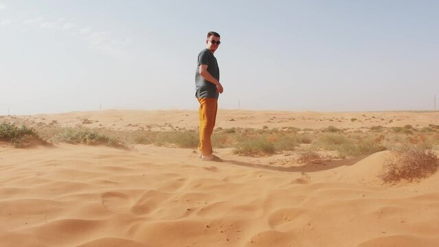 Young man in sunglasses and orange pants stands in the desert among camel thorns and smiles. Slow motion