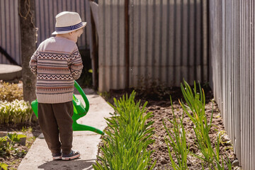 A cute boy in a hat plants and waters seedlings from a watering can in a summer garden, outdoors. The concept of gardening and teaching a child to work. © Юлия Клюева